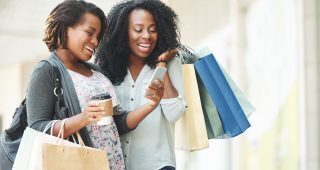 two woman smiling looking down at one of their smartphone's while holding shopping bags and a hot beverage